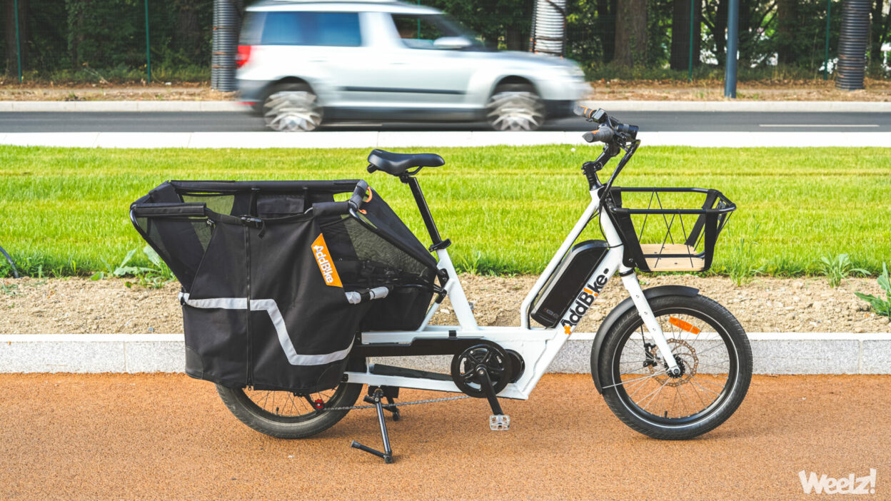 Test vélo longtail AddBike U-Cargo ; Sur cette photo, un véhicule utilitaire et familial du quotidien. A l’arrière-plan, une voiture.